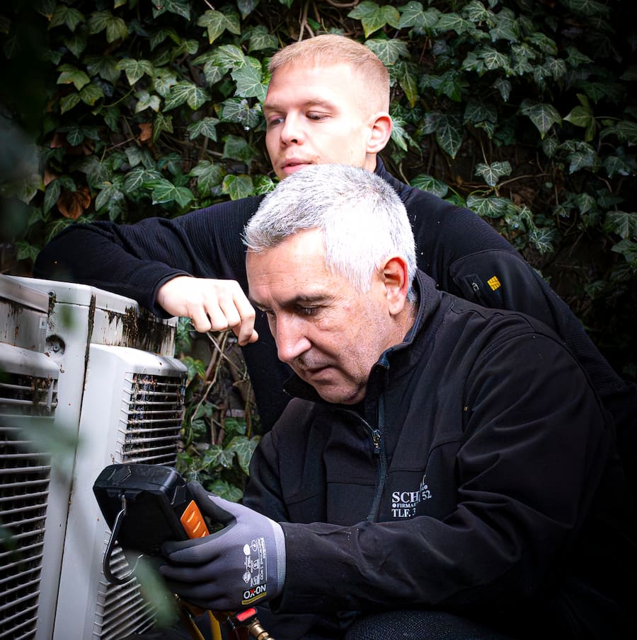 o klimaanlæg-installatører fra Schøtt arbejder sammen om at kontrollere et udendørs klimaanlæg ved hjælp af et digitalt måleinstrument. De står omgivet af grøn beplantning i en gårdhave i København, hvilket illustrerer professionel installation og service af klimaanlæg i byen.